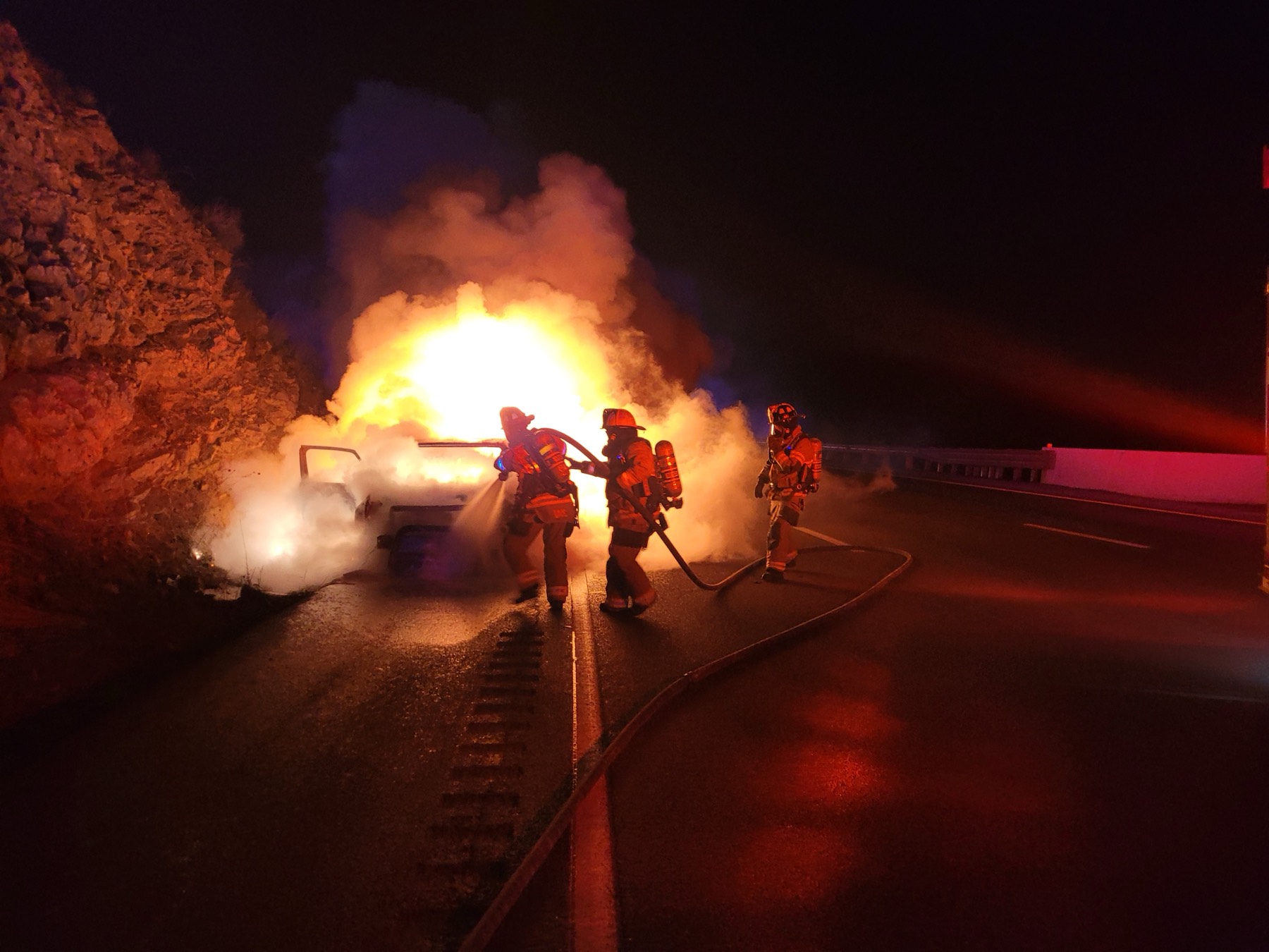 Three firefighters advancing a hose line on a fully involved vehicle fire on I-70 westbound at night.